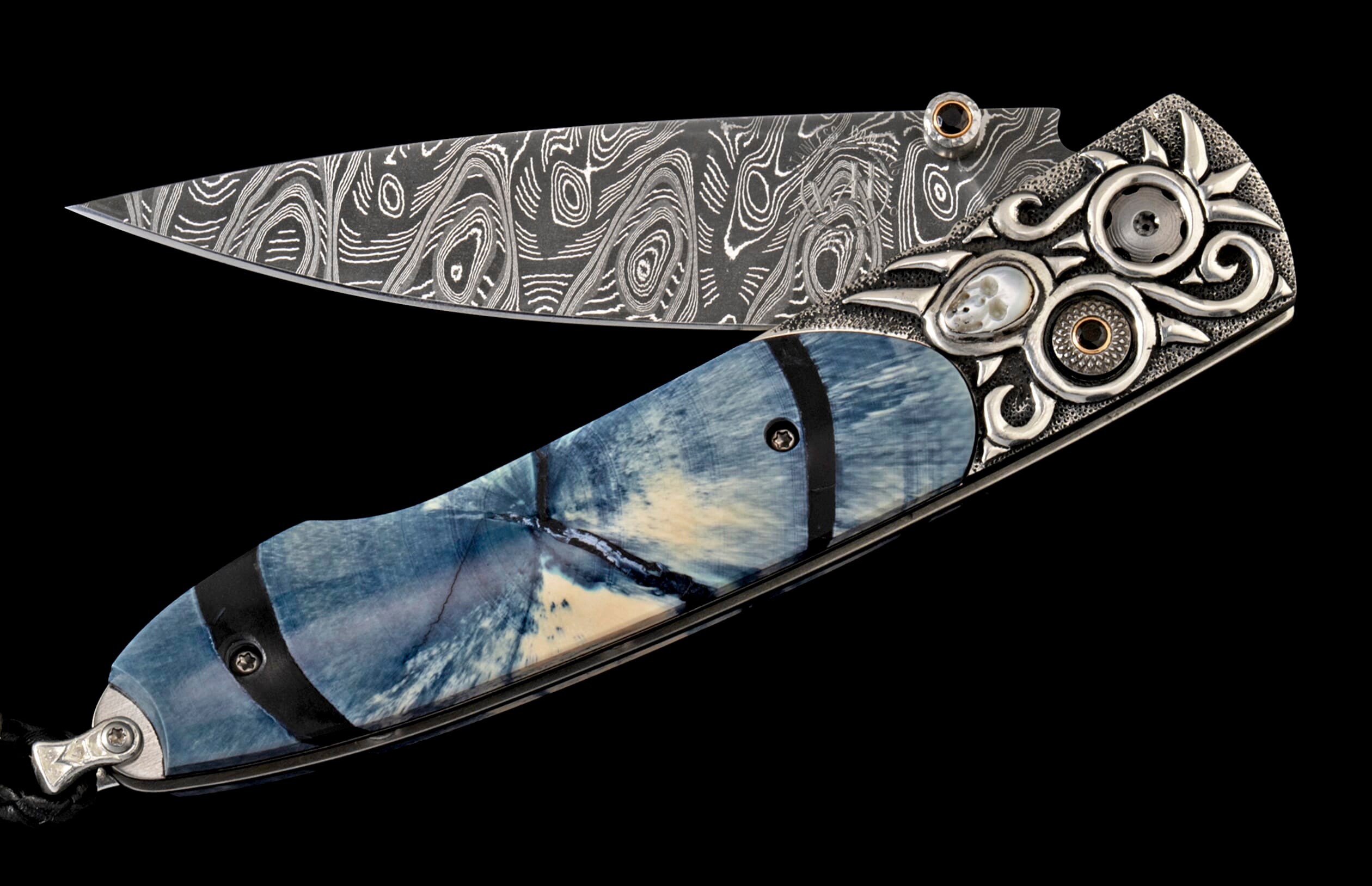 Ornate folding knife with a patterned Damascus steel blade, blue and cream handle, and intricately carved silver bolster featuring skull inlay and black gems.