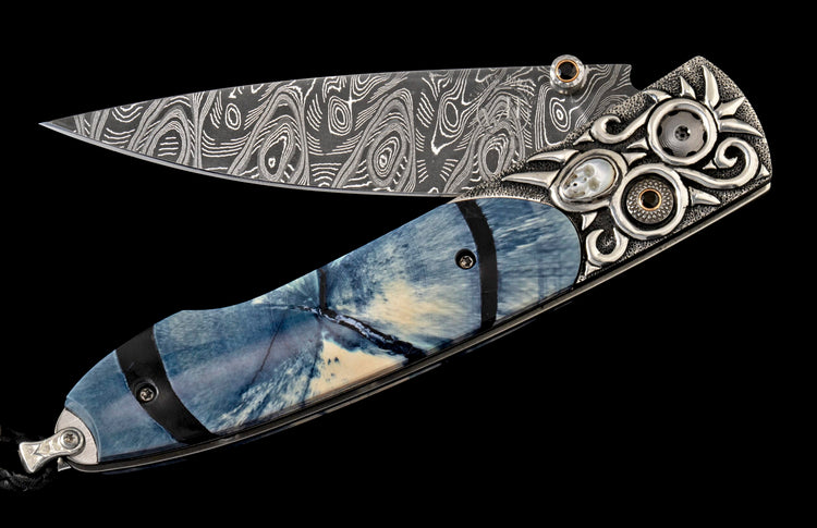 Ornate folding knife with a patterned Damascus steel blade, blue and cream handle, and intricately carved silver bolster featuring skull inlay and black gems.