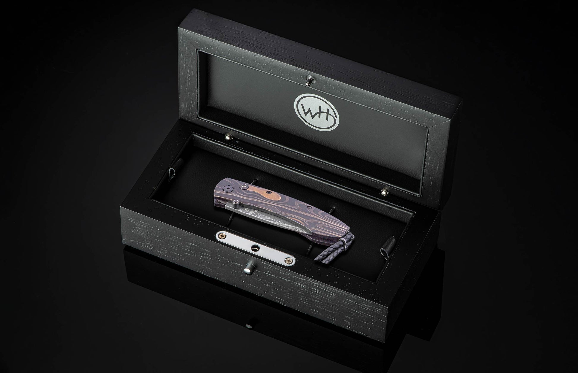 Dark folding knife with purple wood grain handle and Damascus blade by Wharncliffe, displayed in black presentation box with silver hardware, top-down view