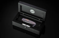 Dark folding knife with purple wood grain handle and Damascus blade by Wharncliffe, displayed in black presentation box with silver hardware, top-down view