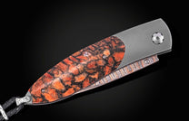 Coral-patterned folding knife with textured orange and black handle, by B05 Ignis, featuring silver hardware and braided lanyard, side view on black background