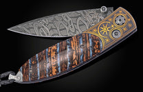 Open folding knife with a patterned Damascus steel blade, a handle of brown and blue fossilized material, and a black metal section with gold star engravings, finished with a braided black leather lanyard.