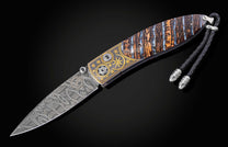 Ornate folding knife with Damascus steel blade, multi-colored Stardust Fossil handle, gold star and sunburst inlays, and a braided leather lanyard.