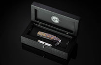Folding knife with gold-patterned bolster and blue-orange striped fossil handle, displayed in an open black wooden box with a 'WH' logo.