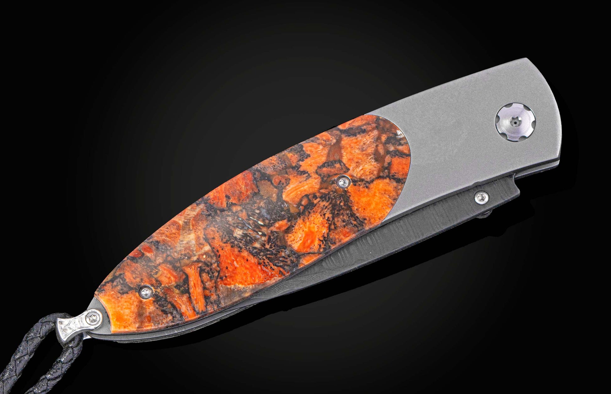 Angled view of a folding knife with an orange and black patterned handle, grey metal frame, Damascus blade, and braided black lanyard.