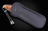 Knife with an orange and brown mottled handle peeking from a black leather sheath with an embossed logo and a braided lanyard.