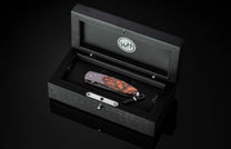 Silver folding knife with a unique orange, brown, and black speckled handle, presented in an open black wooden box with a 'WH' logo and a braided lanyard.