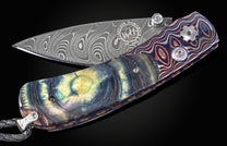 Damascus steel folding knife with a swirling blade pattern, vibrant iridescent handle, and a braided black lanyard.