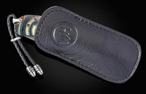 Folding knife with an iridescent blue and yellow handle and Damascus blade, partially in a dark leather sheath with an embossed logo and a braided lanyard.