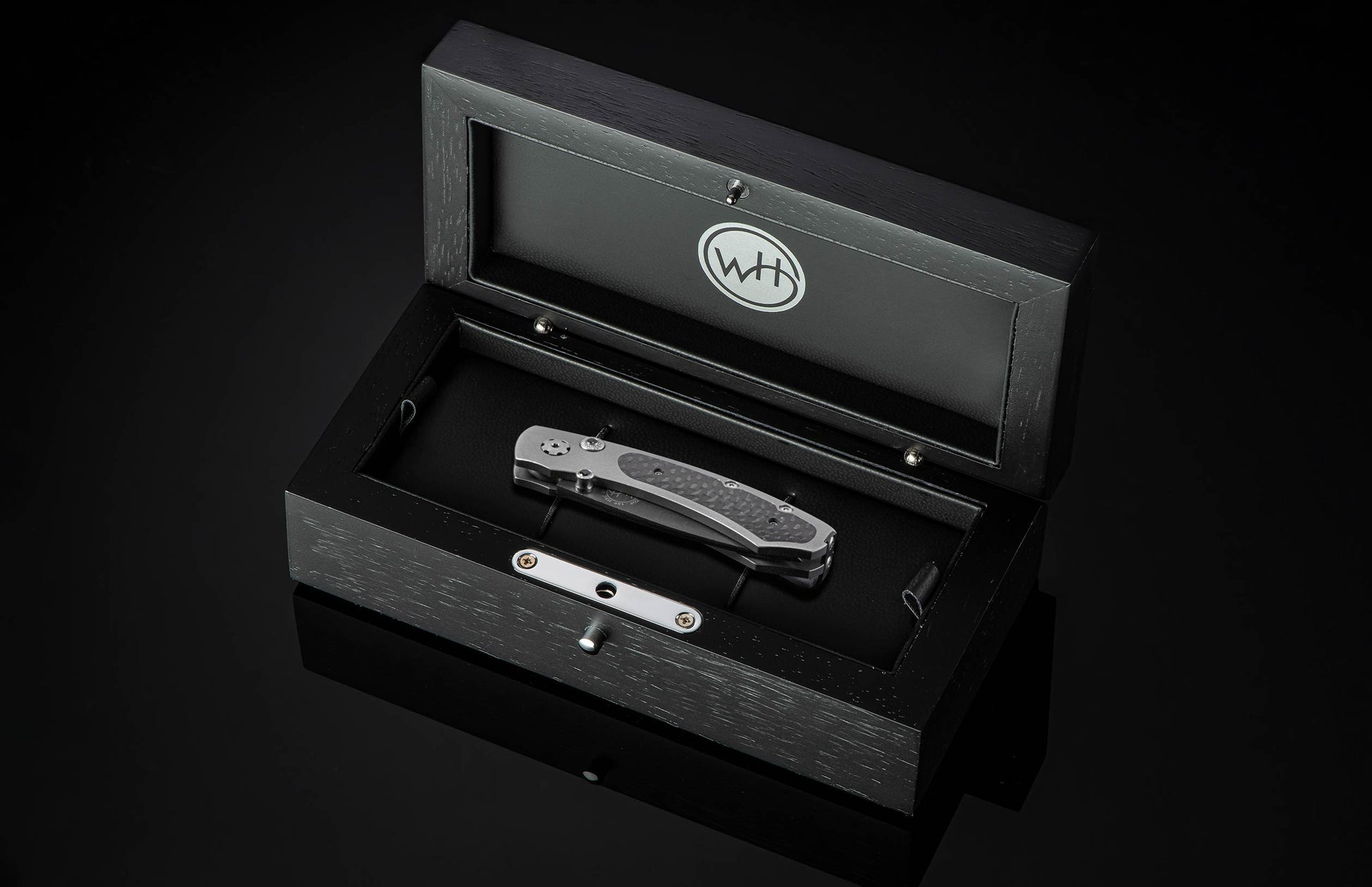 B09 Nightshade folding knife with dark textured handle, resting in an open black wooden box featuring a 'WH' logo.