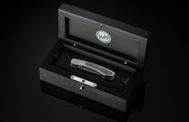 B09 Nightshade folding knife with dark textured handle, resting in an open black wooden box featuring a 'WH' logo.