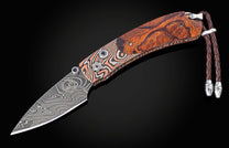 Folding knife with a swirling Damascus steel blade, polished burl wood handle, copper-toned patterned bolster, and a braided brown leather lanyard.
