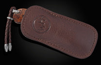 Brown leather knife sheath with a debossed logo and stitching, partially revealing a wooden-handled knife and braided lanyard.