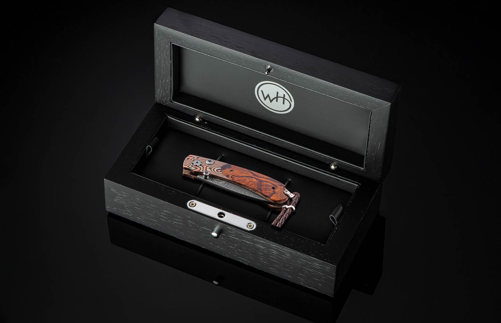 Folding knife with a rich wooden handle and patterned steel blade, resting in an open black presentation box featuring a 'WH' logo.