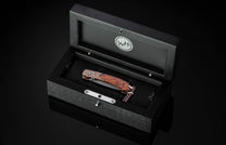 Folding knife with a rich wooden handle and patterned steel blade, resting in an open black presentation box featuring a 'WH' logo.