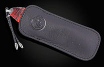 Folding knife with a red patterned handle, partially in a dark leather sheath with an embossed logo and braided lanyard.