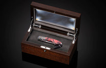 Folding knife with red and black marbled handle and metallic engine-like details, inside an open dark brown wooden display box.