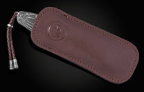 Folding knife with a Damascus-patterned handle and braided brown leather lanyard with silver accents, partially in a brown leather sheath with an embossed logo.