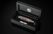 Folding knife with Damascus patterned blade and auburn and grey striped handle, in an open black wooden presentation box with a WH logo.