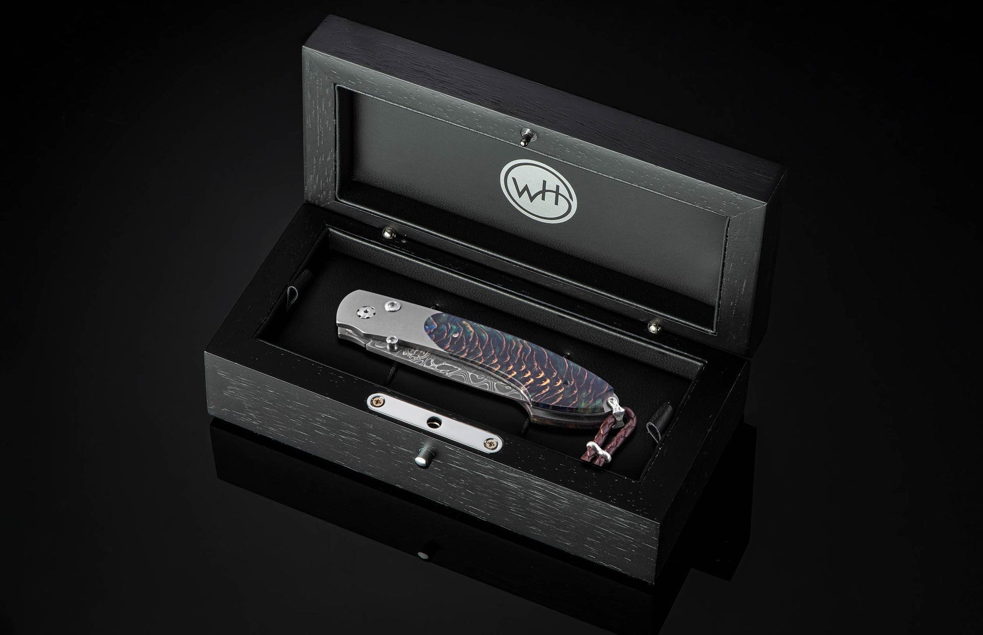 Luxury folding knife with iridescent Aurora Pine handle and damascus blade, by B12, displayed in black textured box with silver hardware, top-down view