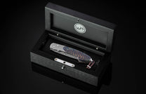 Luxury folding knife with iridescent Aurora Pine handle and damascus blade, by B12, displayed in black textured box with silver hardware, top-down view