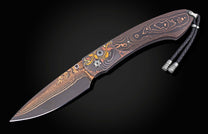 Folding knife with a dark Damascus blade and a handle featuring gold and copper ornate patterns, accented by a braided leather lanyard.