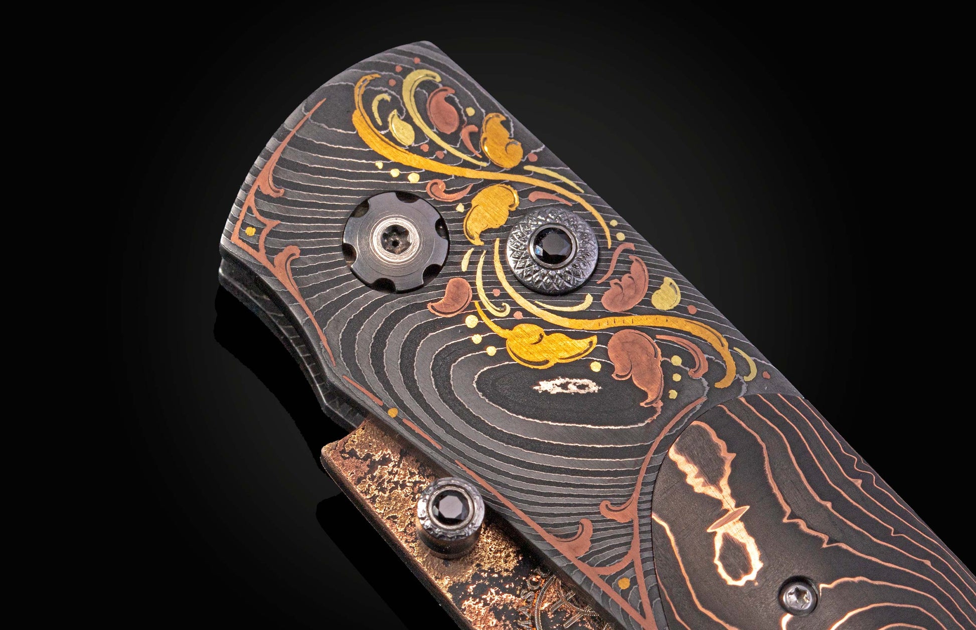 Ornate knife handle with swirling damascus steel, gold and copper leaf inlays, and decorative black jeweled fasteners.