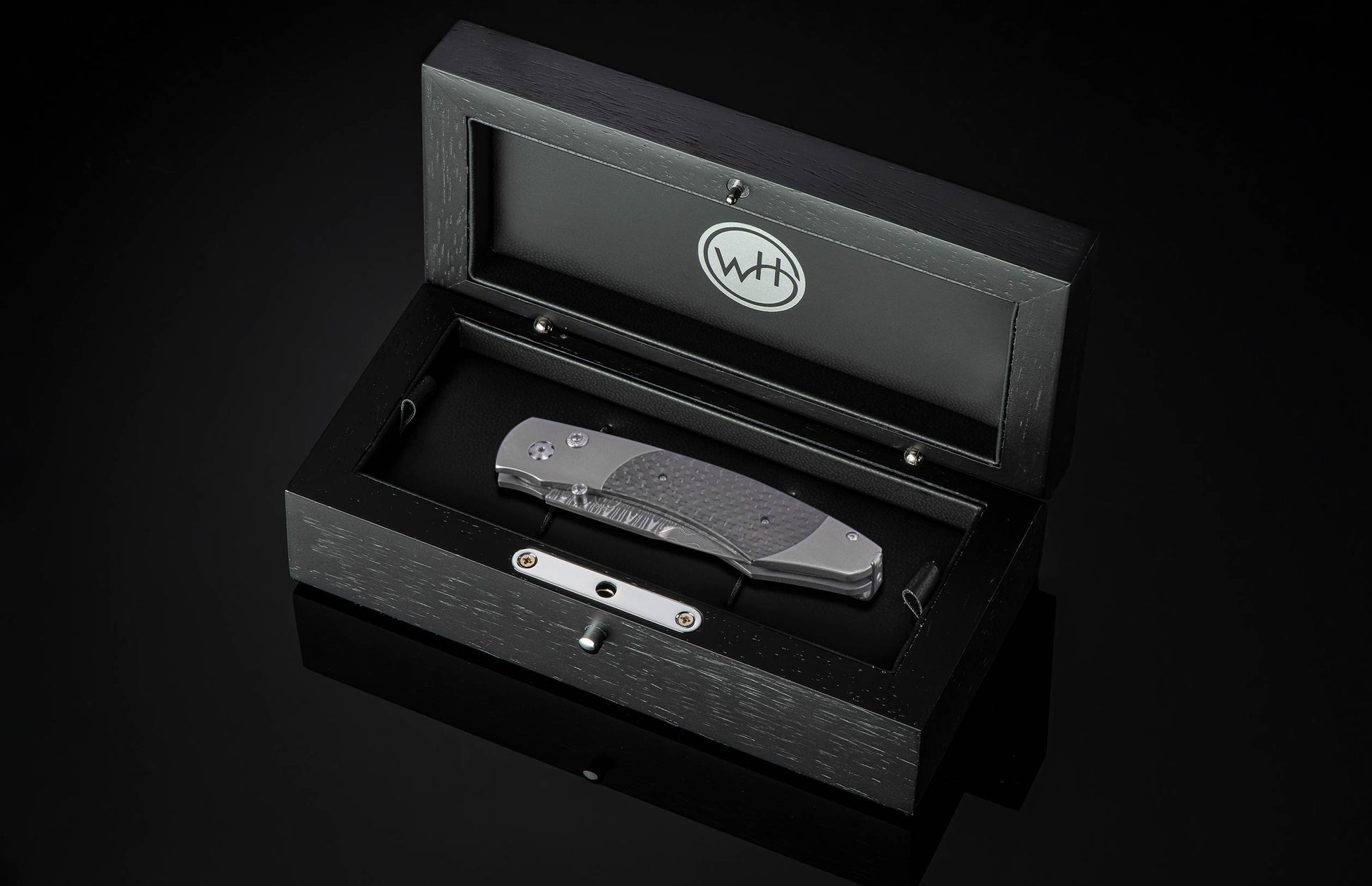Grey folding knife with a textured carbon fiber handle and Damascus blade, displayed inside an open black wooden presentation box with a silver WH logo in the lid.