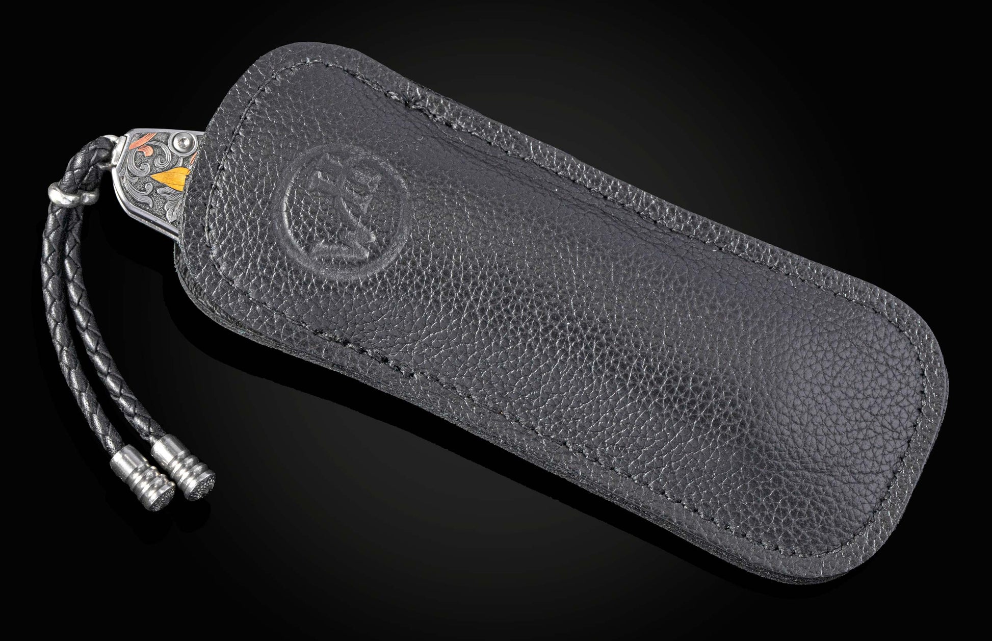 Folding knife with engraved silver-toned handle featuring gold and copper accents, partially sheathed in textured dark leather with an embossed logo, and a braided lanyard.