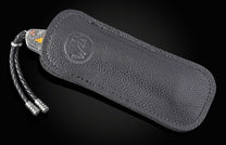 Folding knife with engraved silver-toned handle featuring gold and copper accents, partially sheathed in textured dark leather with an embossed logo, and a braided lanyard.