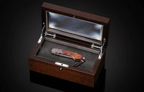 Luxury folding knife with burl wood handle and engraved titanium bolster, displayed in a dark wood presentation box with glass lid.