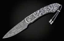 Damascus steel folding knife with black and white mosaic handle by B12, featuring gear embellishments and braided lanyard, side view on dark background
