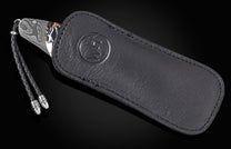 Luxury Damascus steel knife with marbled handle and black leather sheath by B12, featuring braided cord and embossed logo, side view on dark background