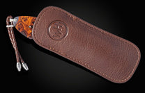 Knife with an orange burl wood handle and braided leather lanyard, partially in a brown pebbled leather sheath with an embossed logo.