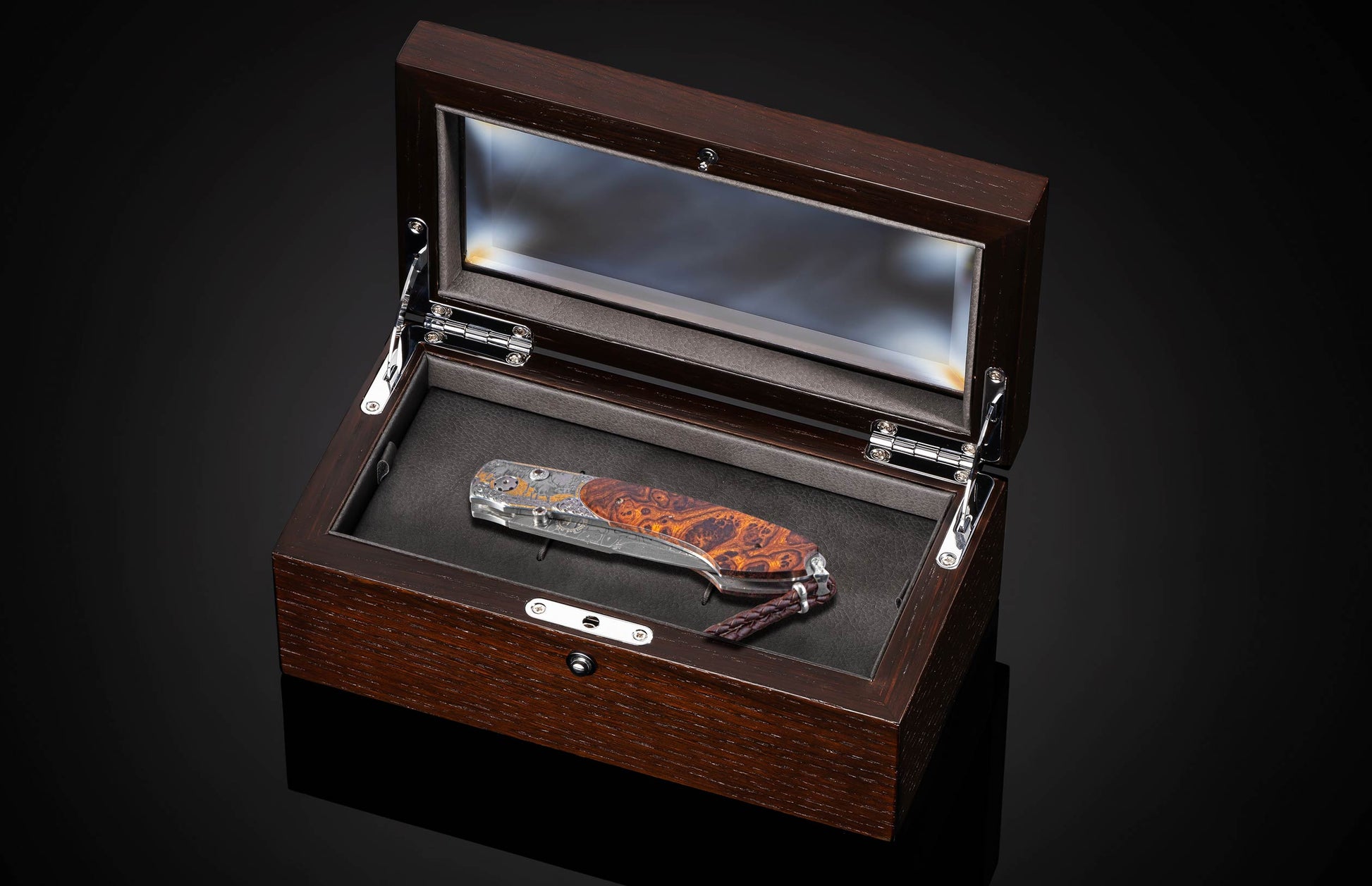 Ornate folding knife with engraved silver and gold accents, a dark orange burl wood handle, and a leather lanyard, presented in an open dark wooden display box.