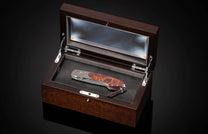 Ornate folding knife with engraved silver and gold accents, a dark orange burl wood handle, and a leather lanyard, presented in an open dark wooden display box.