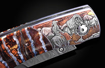 Folding knife with an engraved western frontier scene of a cowboy and covered wagon, alongside a layered brown and blue stone handle and Damascus blade.