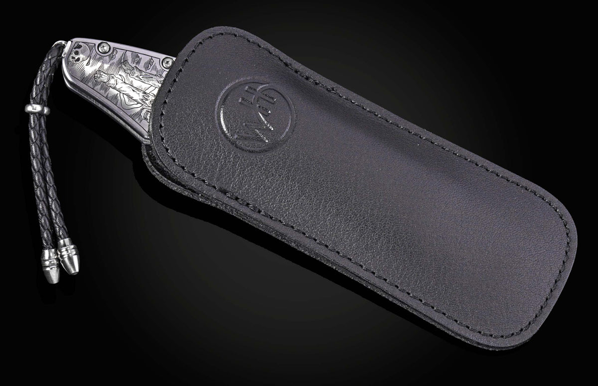 Silver engraved folding knife with Halloween-themed skeleton design, by B12, in black leather sheath with braided cord and metal tips, side view.