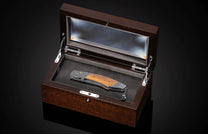 Folding knife with dark wood handle and intricate metal engravings, showcased in an open dark wooden display box.