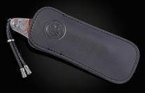 A decorative knife with an intricately engraved silver and copper handle, partly visible from a black leather sheath with a debossed logo and braided lanyard.