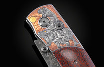 Ornate folding knife with detailed silver raptor engravings on a copper-toned background, yellow eyes, black gemstones, and a reddish-brown dinosaur bone handle.
