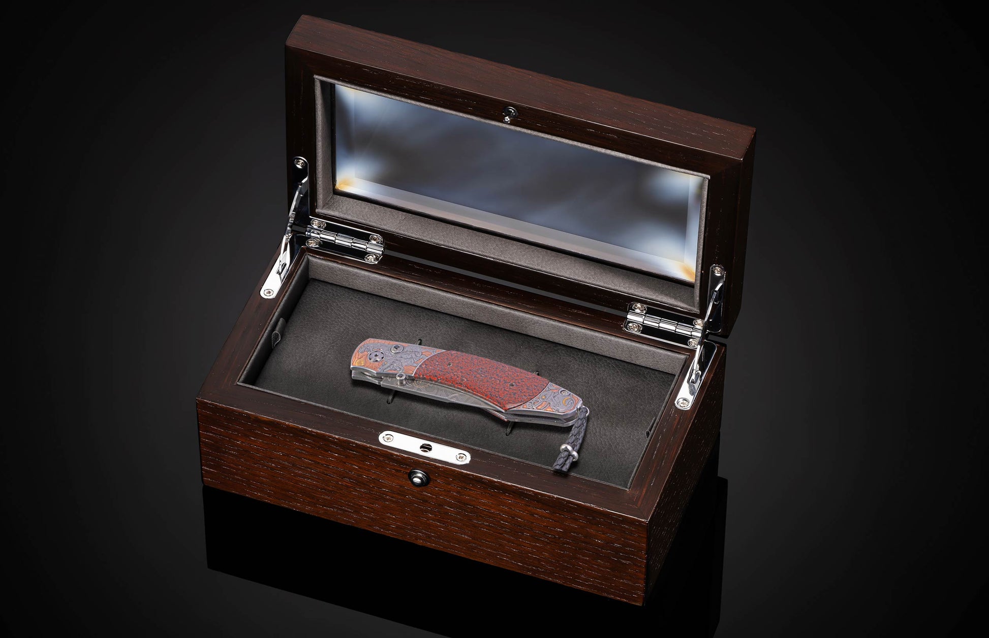 A folding knife with a red, orange, and gray patterned handle and a gray lanyard, displayed in an open dark brown wooden presentation box on a black reflective surface.