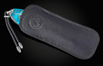 Turquoise handled knife with silver accents, partially in a black pebbled leather sheath featuring an embossed circular logo and braided lanyard.