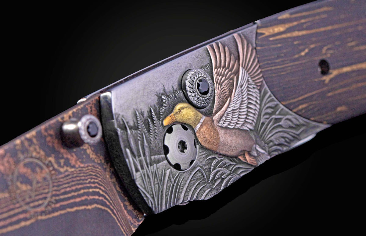 Angled close-up of a knife handle featuring a bronze and silver mallard duck flying over etched reeds, with a black gem and dark wood grain.