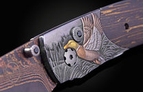 Angled close-up of a knife handle featuring a bronze and silver mallard duck flying over etched reeds, with a black gem and dark wood grain.