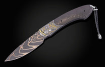 Dark artistic folding knife with patterned Damascus steel blade, intricate gold and silver engraved handle, and braided leather lanyard.