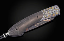 Damascus steel folding knife with an engraved silver and gold skull motif and a braided leather lanyard.