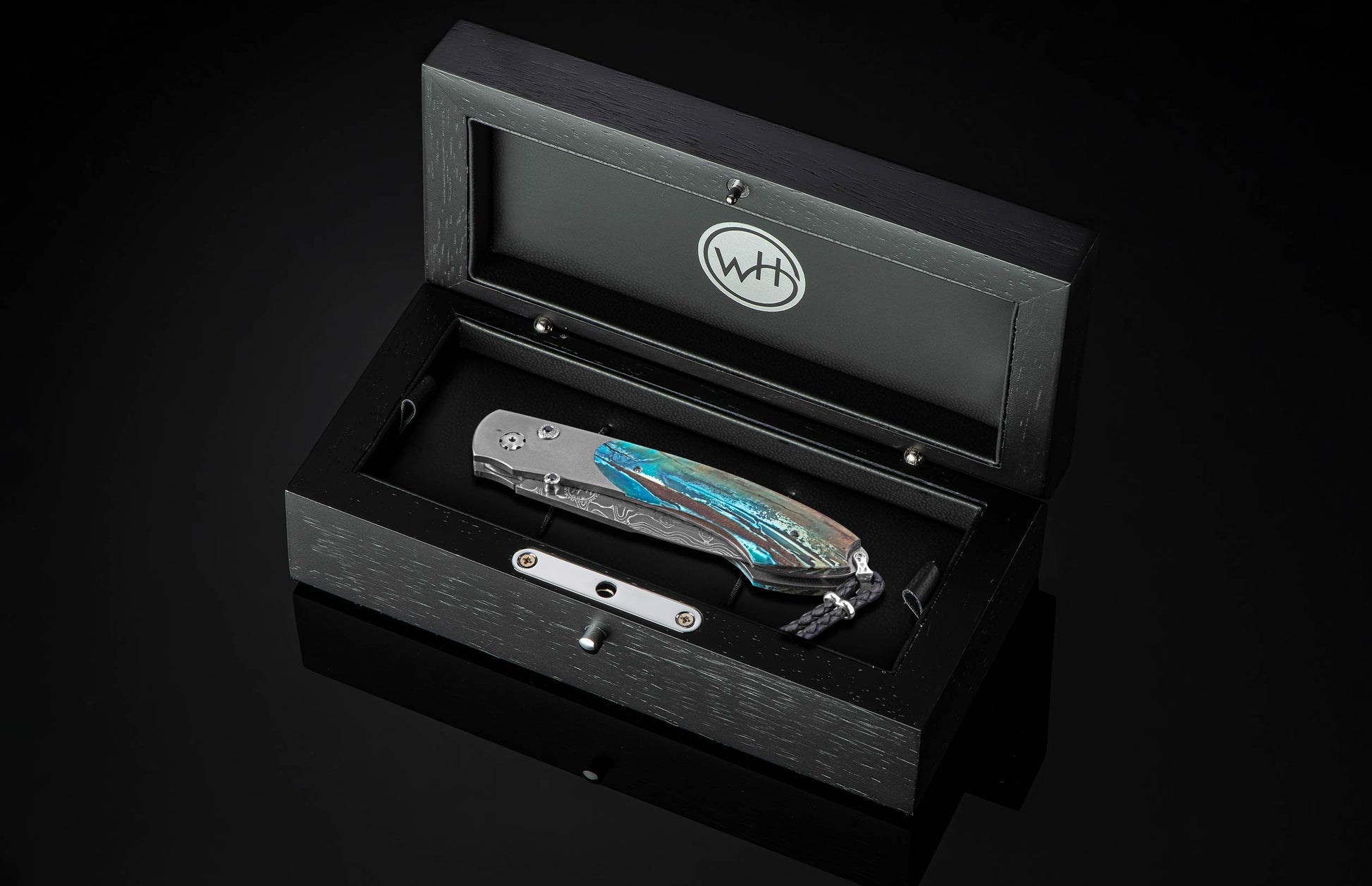 Stainless steel folding knife with turquoise and brown marbled handle by B12, displayed in black wooden box with silver logo, side view