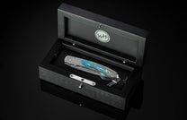 Stainless steel folding knife with turquoise and brown marbled handle by B12, displayed in black wooden box with silver logo, side view