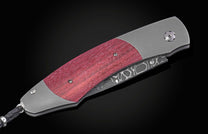 Reddish-purple wood handle folding knife with grey metal frame, featuring a damascus steel blade and a black braided lanyard.